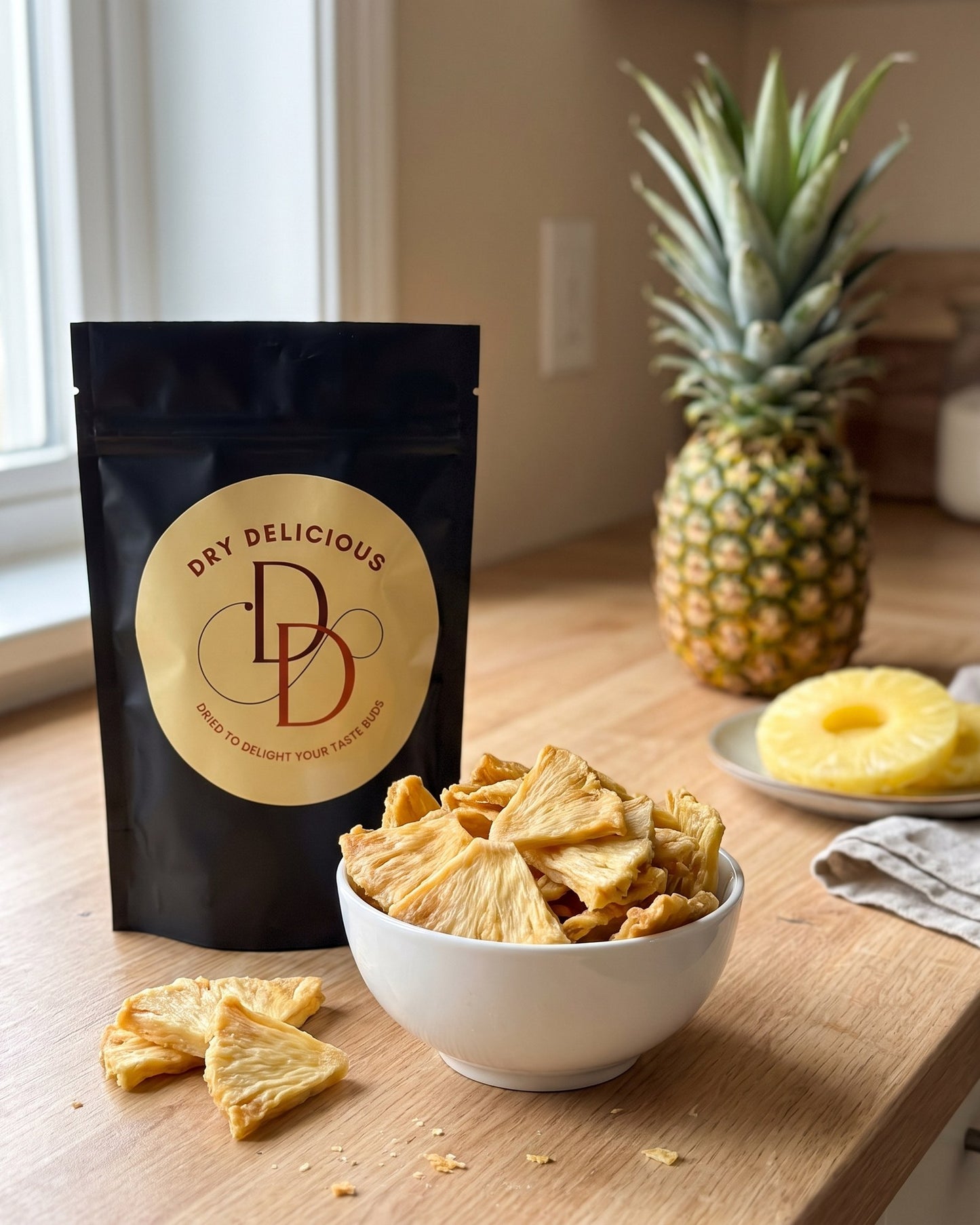 Dry Delicious dehydrated pineapple slices in a black stand-up pouch beside a white bowl of dried pineapple pieces, with a fresh pineapple and pineapple rings on a wooden counter