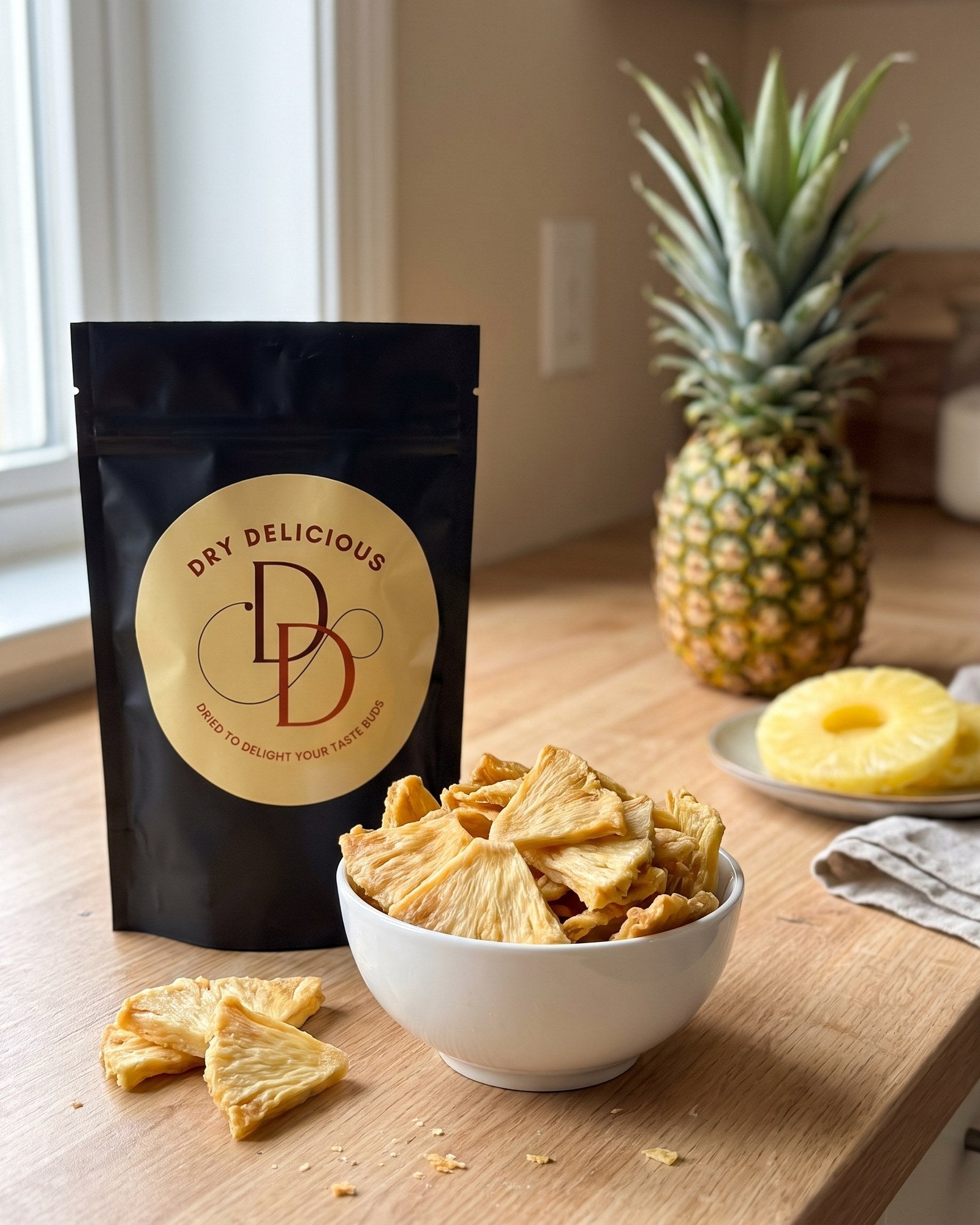 Dry Delicious dehydrated pineapple slices in a black stand-up pouch beside a white bowl of dried pineapple pieces, with a fresh pineapple and pineapple rings on a wooden counter